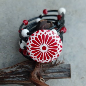 Può includere: Un braccialetto con perline nere e bianche con un bottone in ceramica rosso e bianco. Il bottone ha un design geometrico.