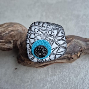 Puede incluir: Broche decorativo de forma cuadrada con superficie estampada en blanco y negro. Presenta un círculo azul claro y un botón negro texturizado en el centro. El broche está sobre un trozo de madera flotante.