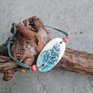 Op de afbeelding: Een tealblauwe en witte keramische bladeren armband met roze kralen en een groen koord.