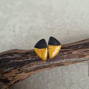 Può includere: Un paio di orecchini a bottone dal design unico. Ogni orecchino ha la forma di un mezzo triangolo, con una parte superiore nera e una parte inferiore gialla con un motivo screpolato. Gli orecchini sono esposti su un pezzo di legno.