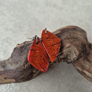 Puede incluir: Un par de pendientes rojos y negros con un patrón texturizado y agrietado. Los pendientes tienen forma de diamante y cuelgan de ganchos de color plateado. Los pendientes se exhiben sobre un trozo de madera flotante.
