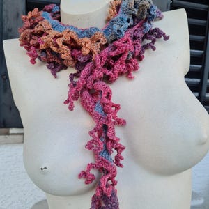 May include: A crocheted scarf in shades of pink, orange, and blue. The scarf is made with a textured stitch and has a long, flowing design.