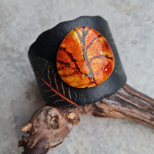 May include: Black leather cuff bracelet with a round, orange and gold resin inlay featuring a tree branch design.