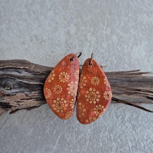 Könnte beinhalten: Ein Paar orangerote Ohrringe mit goldenem Blumenmuster. Die tropfenförmigen Ohrringe haben eine strukturierte Oberfläche. Sie hängen an einem Stück Treibholz vor grauem Hintergrund.