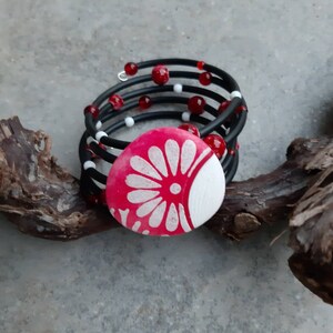 Op de afbeelding: Een zwarte en rode kralen armband met een grote roze en witte bloemvormige knoop. De armband is gemaakt van meerdere strengen zwarte elastische koord met rode en witte kralen.