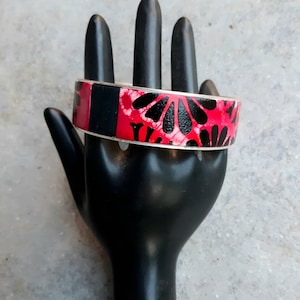May include: A wide silver bangle bracelet with a black and pink floral design. The bracelet is on a black hand model.
