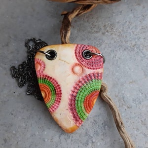 Peut inclure: Collier avec pendentif en forme de médiator de guitare. Le pendentif crème présente un motif circulaire rose, orange et vert. Il est attaché à une chaîne sombre. Bijou unique.