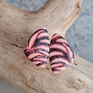 Può includere: Un paio di orecchini triangolari con motivo rosa e nero. Gli orecchini hanno un motivo a spirale di nero e rosa.