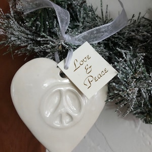 May include: White ceramic heart-shaped ornament with a peace sign embossed on the front. The ornament is hanging from a white ribbon and has a tag that reads "Love & Peace".