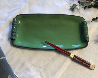 May include: A green ceramic serving dish with raised edges and bamboo-like handles. The dish is on a white tablecloth with a pair of red chopsticks.