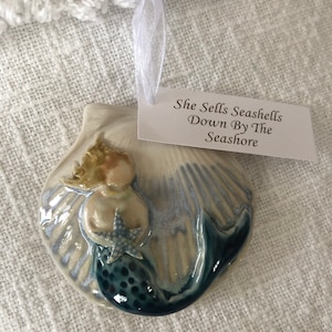 Pode incluir: Um ornamento de concha de cerâmica com uma sereia segurando uma estrela do mar. A sereia tem cabelo loiro e uma cauda azul com manchas azul escuro. A concha é esbranquiçada com detalhes em azul. Uma etiqueta diz "She Sells Seashells Down By The Seashore".