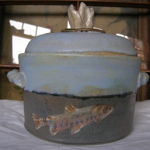 Puede incluir: Una cacerola de cerámica azul y marrón con tapa. La tapa tiene un pomo de cerámica blanca en forma de llama. La cacerola tiene una imagen pintada de una trucha en el lateral.