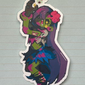 Könnte beinhalten: Ein pixeliger Aufkleber mit einem zombieartigen Charakter mit grüner Haut, lila Haaren und einer rosa Blume. Die Figur trägt ein blaues und rosa Outfit und hat einen kleinen, schwebenden Kopf mit einem Herzen und Sternen.