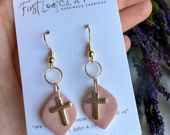 Puede incluir: Pendientes hechos a mano con un colgante de cruz dorada. Los pendientes están sujetos a una base de arcilla rosa en forma de diamante con un patrón texturizado. Los pendientes cuelgan de un gancho dorado. La tarjeta dice "First Love Clay Handmade Earrings".