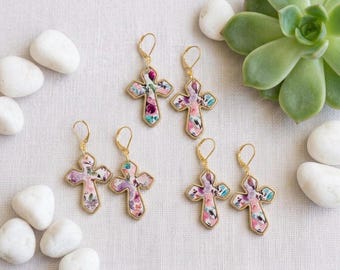 Puede incluir: Tres pares de pendientes colgantes en forma de cruz con un estampado floral en tonos rosa, morado, azul cerceta y blanco. Cada pendiente tiene un borde de metal dorado y un cierre de palanca dorado, dispuestos sobre una tela clara.