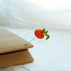 May include: A felt orange pumpkin with a green stem attached to a pin. The pin is on a brown cardboard surface.