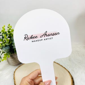 Può includere: Specchio a mano bianco con manico. Lo specchio ha il testo "Rukee Aronson MAKEUP ARTIST" in corsivo nero e una striscia rosa. Lo specchio è tenuto in una mano.