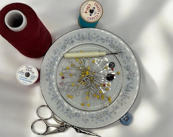 May include: A white ceramic plate with a blue floral pattern, filled with sewing pins, a needle threader, and a few buttons. There are two spools of red thread, one spool of blue thread, and a pair of silver scissors.