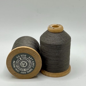 May include: Two spools of gray thread on wooden bobbins. The spools have a label that reads "BELDING CORTICELLI MACHINE TWIST 1000 YARDS". The thread is tightly wound around the wooden spool.