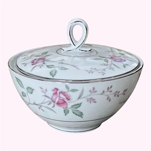 May include: A white porcelain sugar bowl with a lid, featuring a pink rose and green leaf pattern. The rim and handle are silver. The bowl is a classic design for serving sugar or other condiments.