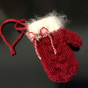 May include: A red knitted mitten ornament with a white cuff and a candy cane ribbon. A small silver bell hangs from the ribbon.