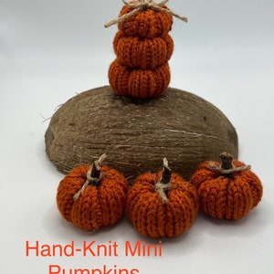 May include: Four hand-knitted orange mini pumpkins, three are laying on a wooden surface and one is stacked on top of itself with a twine bow.
