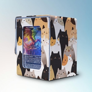 Può includere: Una scatola porta carte con un motivo ripetuto di facce di gatto in vari colori. La scatola ha una finestra trasparente che mostra una carta colorata con un'illustrazione di drago. La scatola è progettata per le carte collezionabili.