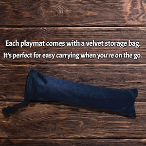 May include: A navy blue velvet storage bag for a playmat, lying on a dark wood surface. The bag is cylindrical with a drawstring closure. Text on the image reads: "Each playmat comes with a velvet storage bag. It's perfect for easy carrying when you're on the go."