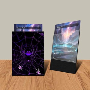 May include: A black card holder with a purple spider web design and the text "Morphic Pool". A card with a landscape scene is displayed in a black stand. The card holder and stand are on a wooden surface.