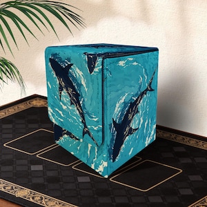 Op de afbeelding: Een kubusvormige opbergkruk met een herhalend patroon van donkerblauwe haaien die in een turquoise oceaan zwemmen. Het ontwerp heeft witte golfdetails. De kruk staat op een zwart oppervlak.