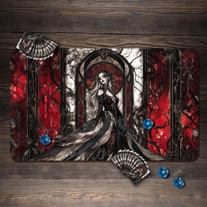 May include: A rectangular playmat with a gothic illustration of a woman in a long black and grey dress, set against a red and white stained-glass window design. Includes blue dice and fan-shaped cards. The mat is on a wooden surface.