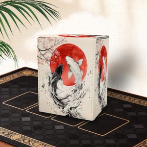 May include: A square, off-white box featuring a Japanese-style illustration of two koi fish swimming in front of a red sun. The design includes cherry blossom branches. The box is on a black surface with a gold border.