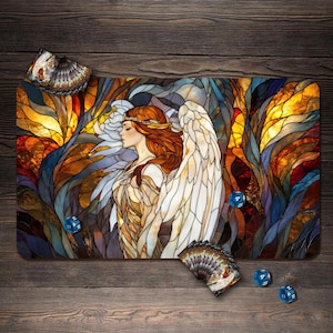 Könnte beinhalten: Eine rechteckige Spielmatte mit einer Illustration im Buntglas-Stil, die eine geflügelte Figur mit langem kastanienbraunem Haar zeigt, umgeben von abstrakten blauen, orangen und gelben Formen. Drei blaue, durchscheinende Würfel liegen darauf.