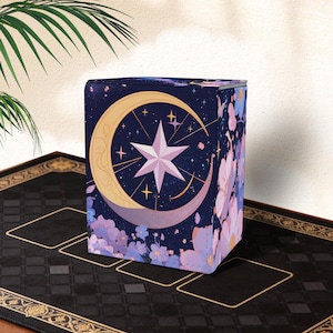 May include: A navy blue card storage box featuring a celestial design. The box has a crescent moon and star design in gold and lavender, with a background of stars and floral accents. The box is sitting on a black mat.