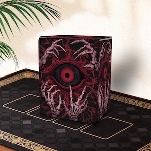 May include: A cube-shaped storage box with a dark red and black design. The box features a central eye motif with a red iris, surrounded by skeletal hands and a web-like pattern. The overall theme is gothic and macabre.