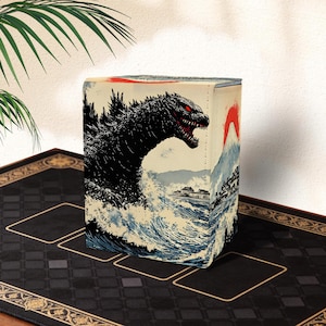 Op de afbeelding: Een kaartenopbergdoos met een Godzilla-ontwerp. De doos toont een zwart-wit illustratie van Godzilla die uit oceaangolven oprijst, met een rode zon en een berg op de achtergrond. De doos staat op een zwarte mat.