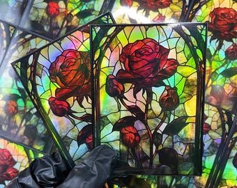 May include: Art cards with a stained glass rose design. The roses are a deep red with green leaves and stems, set against a colourful, iridescent background. The cards are held by a gloved hand.