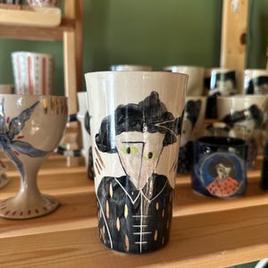 Puede incluir: Un vaso cilíndrico de cerámica con base color crema, que presenta una ilustración pintada a mano de una figura con cabello oscuro, ojos verdes y una prenda negra con cuello, decorada con sutiles acentos verticales dorados y blancos.