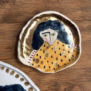 Op de afbeelding: Handbeschilderde keramische schaal met een gouden rand en een vrouwenportret. De vrouw heeft donker haar, groene ogen en draagt een gele jurk met zwarte en gouden accenten. De schaal heeft een onregelmatige vorm.
