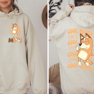 May include: A beige hoodie with a cartoon dog on the front. The dog is wearing a red collar and has a white background. The text "MUM" is below the dog. The back of the hoodie has the text "COOL MOMS CLUB" repeated in a pattern.