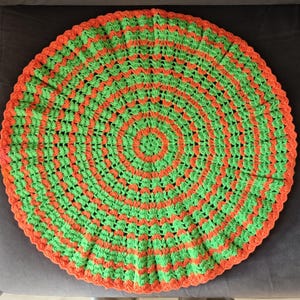 Peut inclure: Nappe ronde faite à la main au crochet dans une palette de couleurs vives. Le motif présente des cercles concentriques de vert et d'orange alternés, avec une bordure orange. La nappe est posée sur une surface grise.