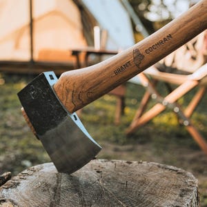 May include: A wooden-handled axe with a black metal head. The handle is engraved with the text "BRANT COCHRAN". The axe is resting on a wooden stump.
