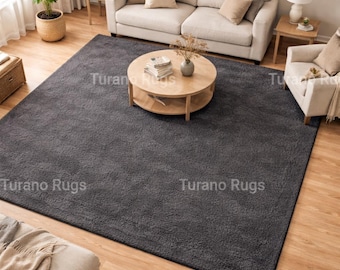 May include: A large, charcoal gray rug dominates a living room setting. The rug is rectangular and covers a significant portion of the wooden floor. The room features a light-colored sofa, armchairs, and a round coffee table, creating a cozy atmosphere.