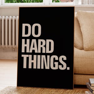 May include: A framed black poster with the words "DO HARD THINGS" in bold white letters. The poster is propped up on a rug in front of a window and a beige couch. A potted plant and a stack of books are also visible.