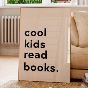 May include: A framed print with the words "cool kids read books" in black text. The print is leaning against a beige sofa, with a window and a potted plant in the background. The room has a neutral color palette.