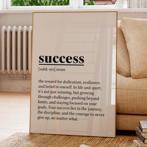 May include: A framed print with the word "success" in a bold, black font. The definition below describes success as the reward for dedication and resilience. The print is displayed in a well-lit room with a window and a beige sofa.