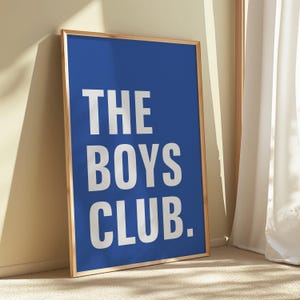 May include: A framed poster with a royal blue background and the white text "THE BOYS CLUB." The poster is in a light brown wooden frame and leans against a wall. The room has a neutral color palette.