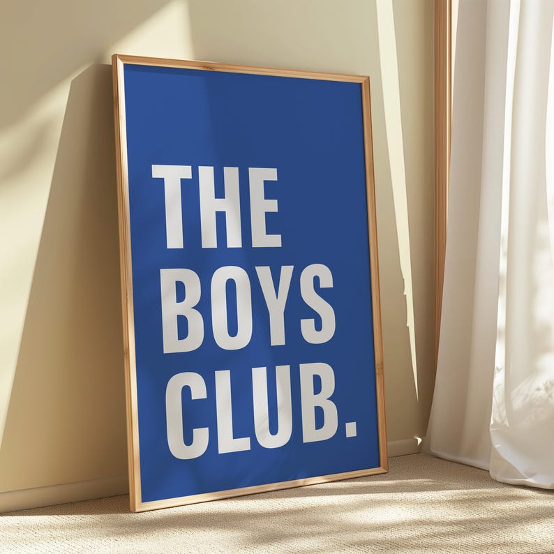 May include: A framed poster with a royal blue background and the white text "THE BOYS CLUB." The poster is in a light brown wooden frame and leans against a wall. The room has a neutral color palette.