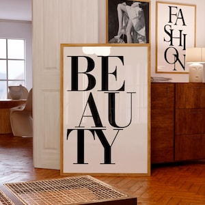 Puede incluir: Un estampado enmarcado con la palabra "BEAUTY" en letras mayúsculas negras grandes. El estampado está en una habitación con otras obras de arte enmarcadas y muebles de madera. La habitación tiene un ambiente cálido.