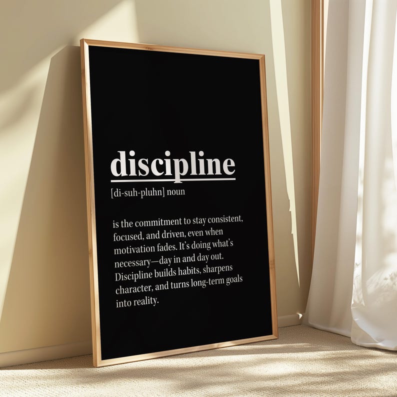 May include: A framed black and white print with the definition of "discipline." The word "discipline" is in a bold, white font at the top, followed by its pronunciation and definition. The print is in a gold frame.